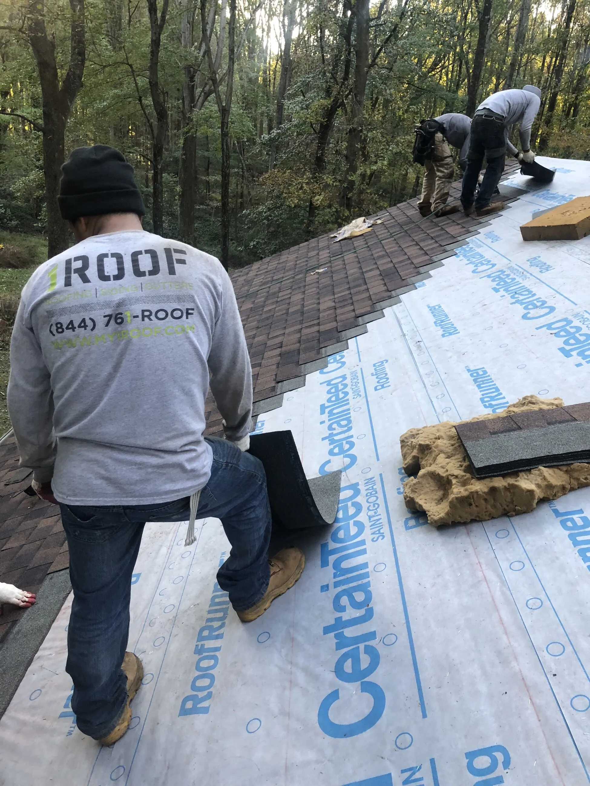 1ROOF crew laying underlayment on a roof deck surrounded by woods