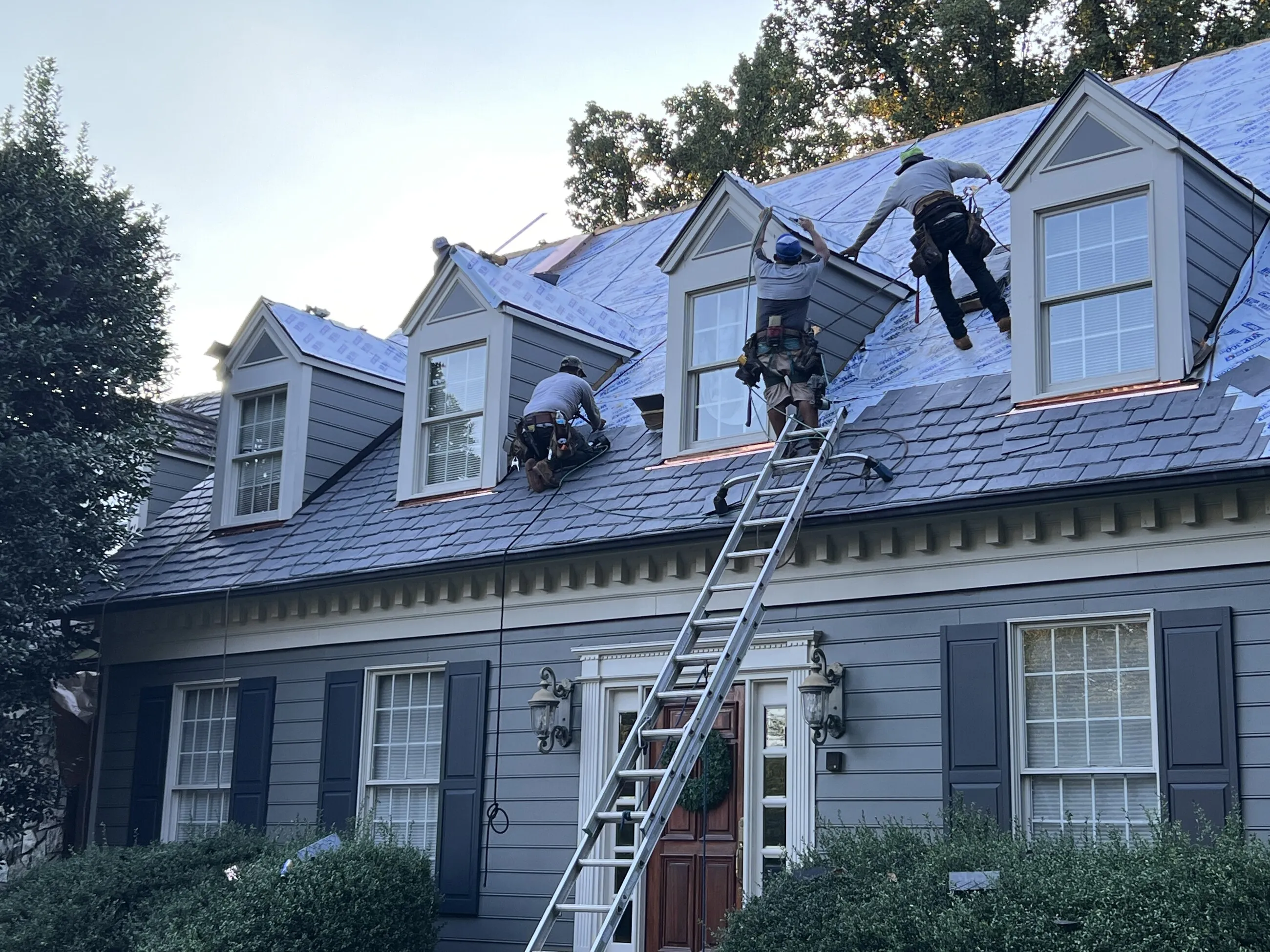1ROOF crew working across the dormers of a gray colonial during a roof replacement
