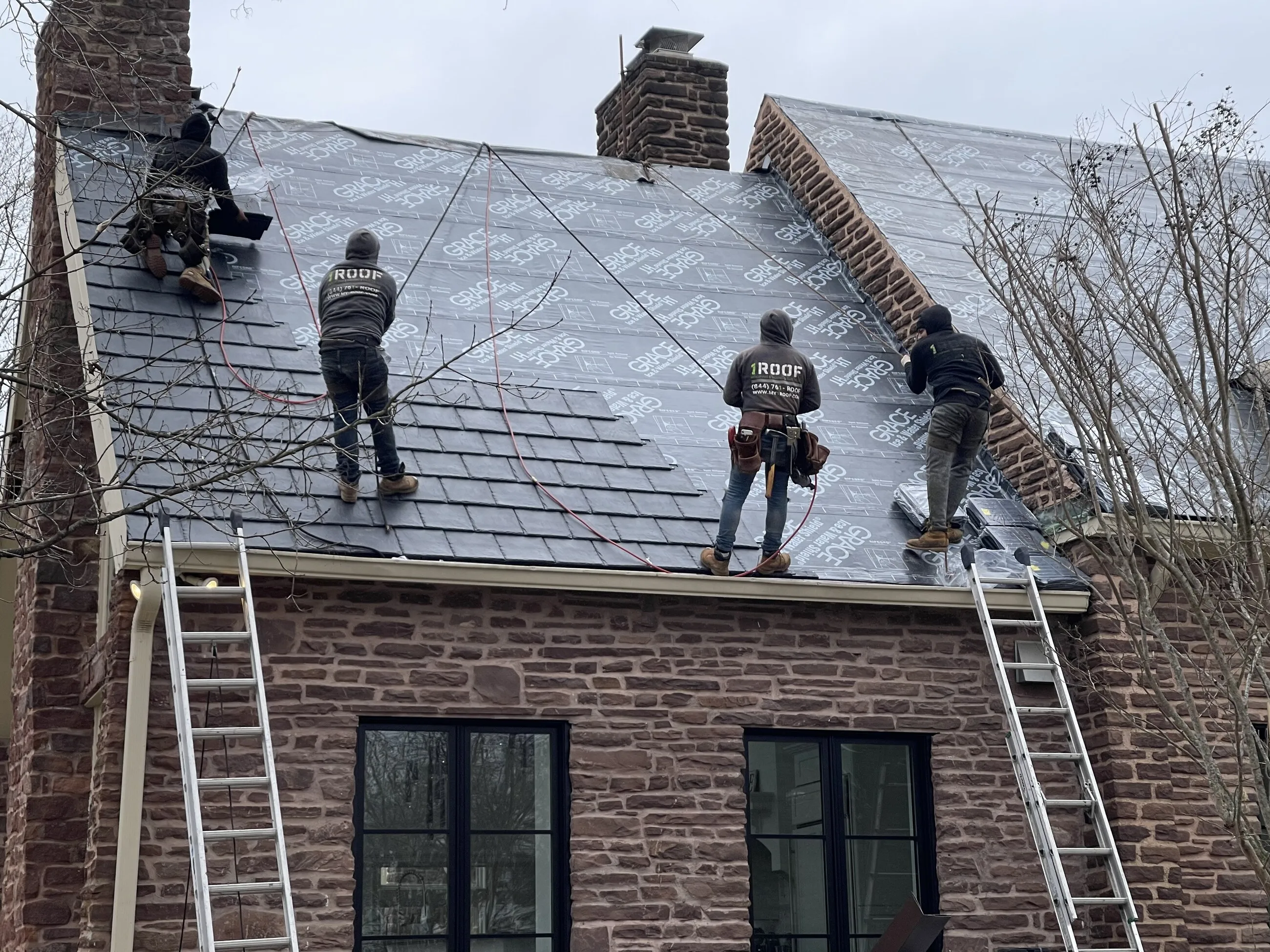 1ROOF crew installing underlayment on the steep roof of a stone home